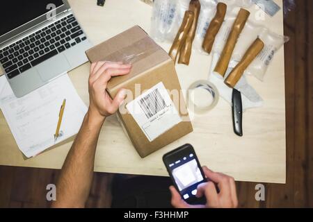 Erhöhte Ansicht der jungen mans Hände mit Smartphone Scannen Lieferung Etikett auf Verpackung Stockfoto