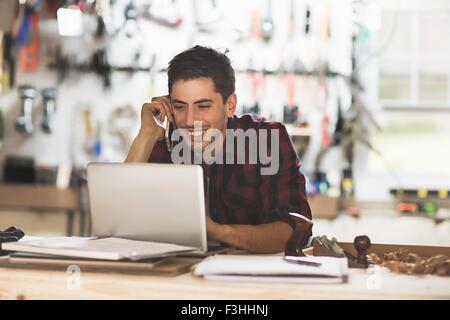 Junger Mann sitzt am Schreibtisch im Workshop am Telefon mit Laptop lächelnd Stockfoto