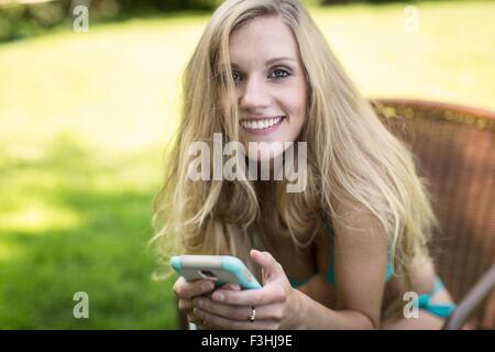 Porträt der jungen Frau mit Bikini mit Smartphone im Garten Stockfoto