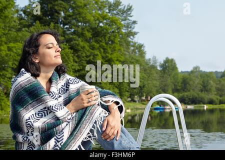 Frau eingewickelt in Decke sitzen am Seeufer, New Milford, Pennsylvania, USA Stockfoto