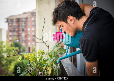 Attraktive junge Mann auf Balkon Bewässerung von Pflanzen im Feld von blauen Gießkanne an sonnigen Tag mit Feld im Hintergrund Stockfoto