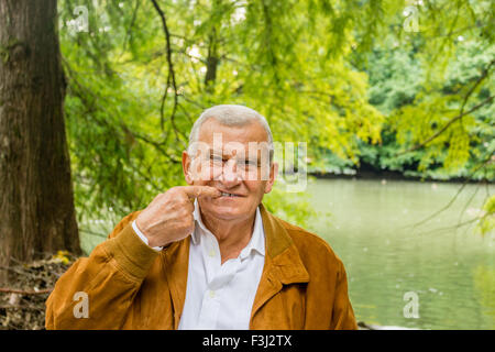 Senior mit Wildleder Jacke und weißes Hemd in einem grünen Park zeigt Zahnersatz Stockfoto