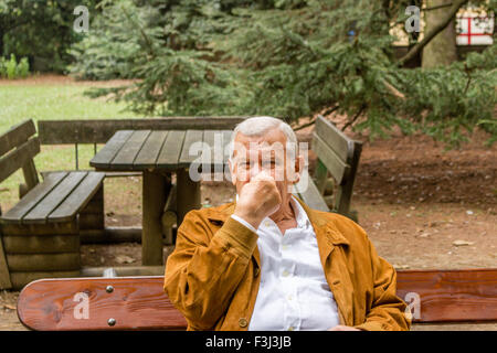 Senior mit Wildleder Jacke und weißes Hemd in einem grünen Park, Verstopfung der Nase Stockfoto