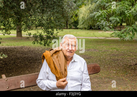 Senior mit Wildlederjacke über Schulter und weißen Hemd in einem grünen park Stockfoto