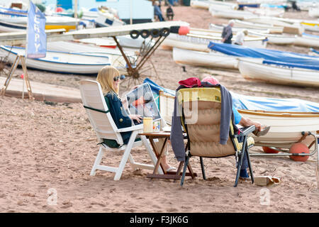 Mann und Frau sitzen in Liegestühlen umgeben von Boote am hinteren Strand Teignmouth Devon England Bootfahren Zeitschriften lesen Stockfoto