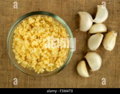 Aerial Makro fein gehackten Knoblauch in einer Schüssel gegen Gunny Tuch mit ganzen Knoblauchzehen im Hintergrund Stockfoto