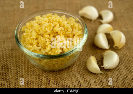 Nahaufnahme des fein gehackten Knoblauch in eine Glasschüssel mit Knoblauchzehen im Hintergrund gegen einen Sack Stoff Textur. Stockfoto