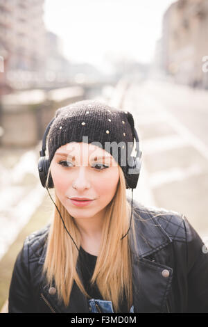 Halbe Länge der junge schöne blonde glatte Haare Frau in der Stadt mit Kopfhörern Musik hören, mit Blick auf richtige, nachdenklich - ernst, nachdenklich, entspannend, Technologie-Konzept Stockfoto
