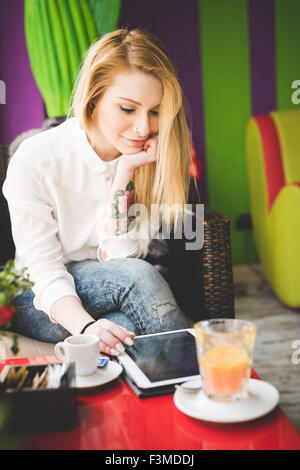 junge schöne blonde glatte Haare Frau in der Stadt sitzen im Restaurant mit Tablet online, verbunden, auf der Suche nach unten und tippen auf den Bildschirm - Technik, soziale Netzwerk-Konzept Stockfoto