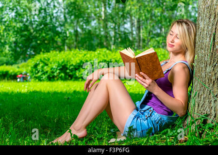 attraktive junge Frau, die einen Roman im Park in der Nähe eines Baumes sitzen lesen Stockfoto