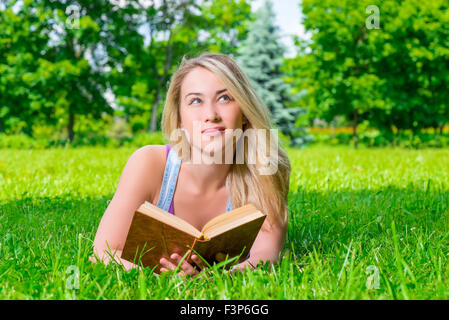 hübsche Blondine auf der Wiese liegen und hält in Händen dickes Buch Stockfoto