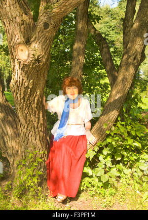 Europäerin mit braunem Schuss Haar in weißer Bluse und roten Rock im Park sitzen auf großen Baum. Stockfoto