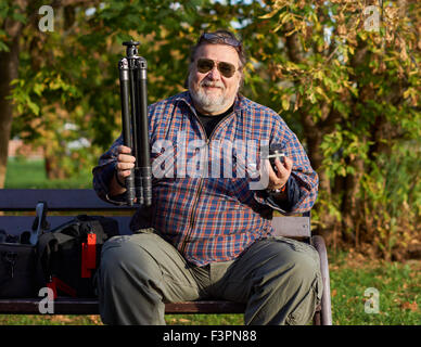 Glücklicher Mensch in Sonnenbrille sitzt auf der Bank im Park mit Stativ in der einen Hand und Kugelkopf in der anderen Stockfoto