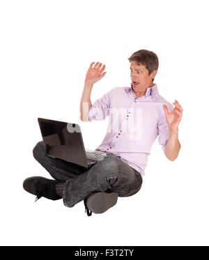 Ein junger Mann, auf dem Boden mit seinem Laptop auf dem Schoß sitzen und hat Angst was He sehen auf dem Laptop isolierten Feind weiß Stockfoto