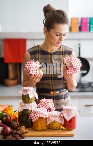 Eine Frau steht mit Gläsern von Gemüse, die sie gerade erhalten, hat. Auf einem Holzbrett auf dem Küchentisch sind weitere Gläser von Gemüsekonserven bereit. Stockfoto