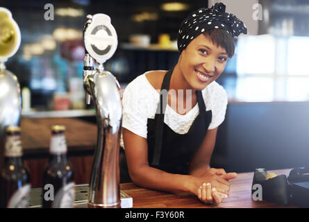Attraktive junge afroamerikanische Kleinunternehmer gekleidet in ein Kopftuch und Schürze gelehnt auf dem Tresen der ihr Pub smili