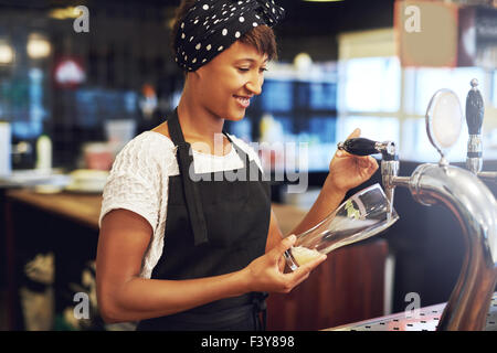 Junge attraktive weibliche afrikanische amerikanische Barkeeper gießen ein Glas Bier vom Fass aus einem Hahn hinter th Abgabe Stockfoto