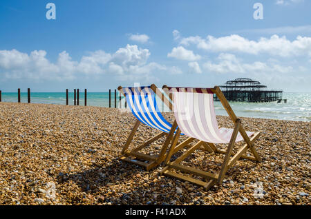 Zwei Liegestühle auf dem einzigen Strand in Brighton und Hove mit den Resten der West Pier in der Ferne, East Sussex, England. Stockfoto