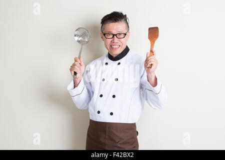 Porträt von zuversichtlich 50er Jahre reife asiatische männlichen Chef in einheitlichen Händen halten Küchenhelfer und lächelnd, stehend auf einfache Zeitmessung Stockfoto