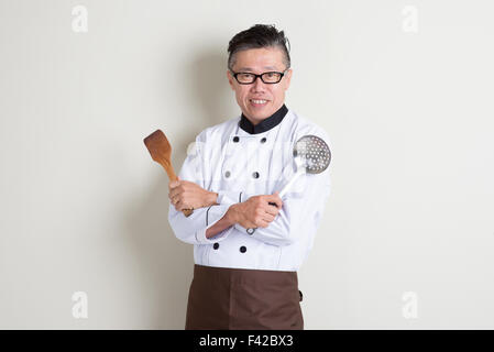 Porträt von zuversichtlich 50er Jahre reife asiatische männlichen Chef in einheitlichen Hände halten Küchengeräte, stehend auf einfarbigen Hintergrund mit shado Stockfoto