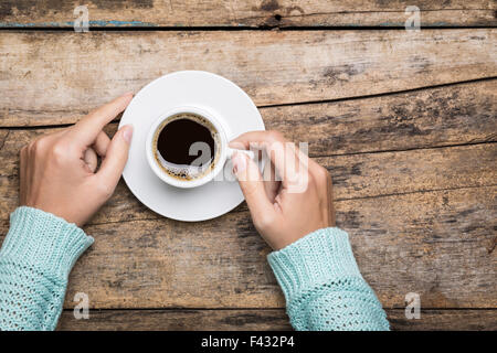 Frauenhand in Pullover halten eine Tasse starken Kaffee auf Holztisch. Kaffee-Fan Draufsicht Hintergrund mit Textfreiraum Stockfoto