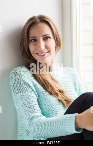 Schöne junge lächelnde Frau sitzt auf der Fensterbank. Ziemlich kaukasische Mädchen auf der Suche in einer Kamera Stockfoto