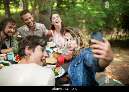 Ein Picknick im Wald. Freunde sitzen um einen Tisch, eine Frau, die eine selfy der Gruppe. Stockfoto