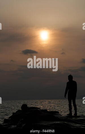 Silhouette des Menschen auf Felsen über dem Meer die Sonne niedrig am Himmel hinter Wolke Dunst Stockfoto