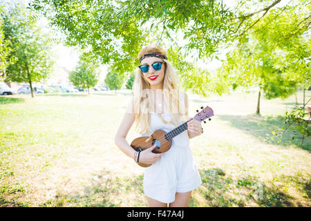 Knie Figur des jungen schönen kaukasischen langes blondes glattes Haar Hippie Frau posiert in einem Stadtpark, eine Ukulele zu spielen, auf der Suche Stockfoto