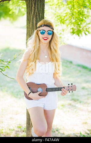 Knie Figur des jungen schönen kaukasischen langes blondes glattes Haar Hippie Frau posiert lehnte sich gegen einen Baum in einem Stadtpark, eine Ukulele zu spielen, suchen Sie in der Kamera, lächelnd - Musik, Inspiration Konzept Stockfoto