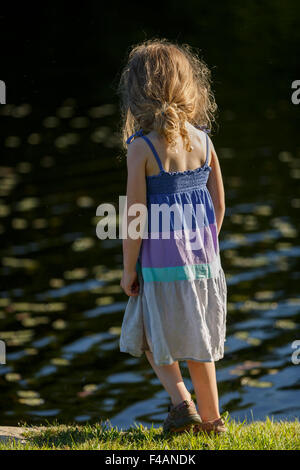 Junges Mädchen im Sommer Kleid mit offenen langen Haaren steht mit Rücken zur Kamera auf dem Rasen am Gewässerrand Stockfoto