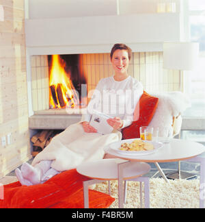 Frau im Liegestuhl am offenen Kamin mit einem Buch in der hand. Stockfoto