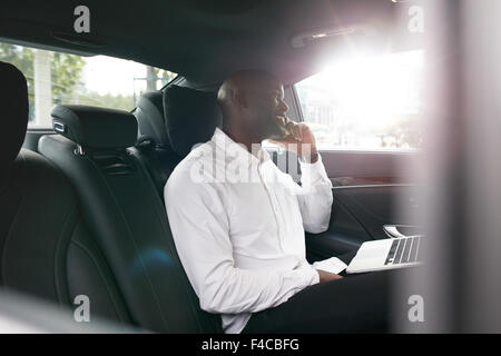 Afrikanischen Geschäftsmann mit Laptop telefonieren mit Handy in einem Auto. Junge Unternehmer arbeiten während der Fahrt zum Büro in Stockfoto