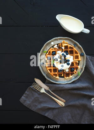 Weiche belgische Waffeln mit Heidelbeeren, Honig und Schlagsahne auf Vintage Metallplatte über schwarzem Holz, Draufsicht, co Stockfoto