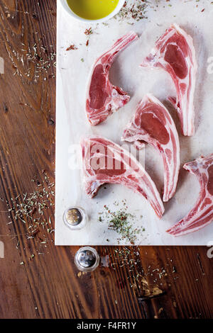 Rohe Lammkoteletts mit Salz, Pfeffer, getrocknete Kräuter und Schüssel mit Olivenöl auf Marmor Brett über Holztisch. Ansicht von oben. Stockfoto