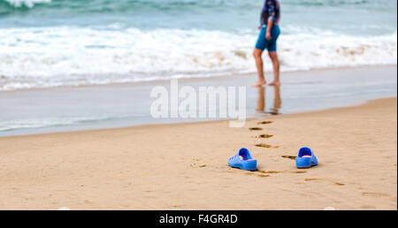 Strand-Hausschuhe in den Vordergrund und verschwommene Silhouette einer Frau in Wellen Stockfoto