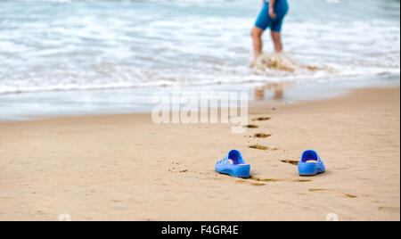 Strand-Hausschuhe in den Vordergrund und verschwommene Silhouette einer Frau in Wellen Stockfoto