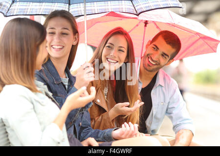 Vier Freunde reden im Freien an einem regnerischen Tag unter Sonnenschirmen wartet in einem Bahnhof Stockfoto