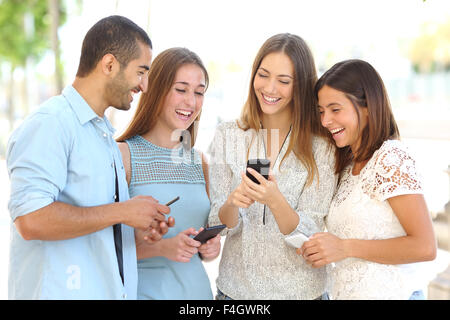 Vier Freunde lachen glücklich und beobachtete soziale Medien in ein Smartphone in der Straße jeder mit seinem eigenen Handy Stockfoto