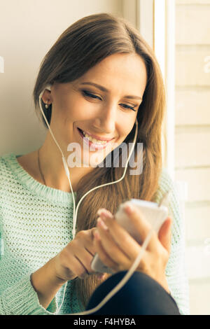 Lächelnde Mädchen Spaß mit Musik hören oder mit ihrem Smartphone. Glückliches schönes Mädchen entspannen Sie am Fensterbrett. Warm Stockfoto