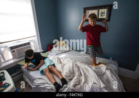 Ein kleiner Junge schaut auf seinen Tablet, während sein jüngere Bruder ärgerlich auf seinem Bett tanzt Stockfoto