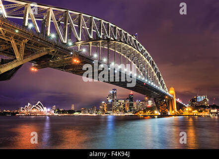 Ostseite der Sydney Harbour Bridge bei Sonnenuntergang mit heller Beleuchtung der Stahlbogen und Spalten in den unscharfen Gewässern reflektieren Stockfoto