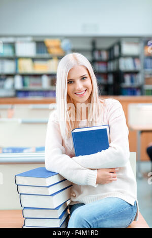 Porträt einer lächelnden jungen Frau sitzt auf dem Schreibtisch mit Stapel Bücher in der Universitätsbibliothek und Blick in die Kamera Stockfoto