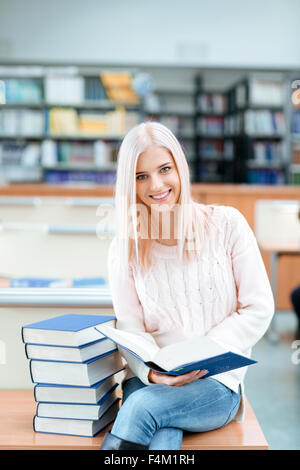 Porträt von einem glücklichen Mädchen sitzt auf dem Schreibtisch mit Stapel Bücher in der Universitätsbibliothek und Blick in die Kamera Stockfoto