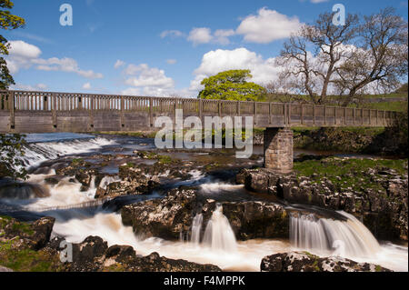 Fluß Wharfe fließt (slow-Shutter Geschwindigkeitsansicht) unter Fußgängerbrücke & Blau Himmel - sonnige malerische Linton Falls Wasserfall, Grassington, Yorkshire, England, UK. Stockfoto