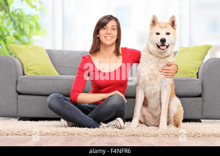 Junge Frau sitzt auf dem Boden mit ihrem Akita Hund und schaut in die Kamera vor einem grauen Sofa zu Hause Stockfoto