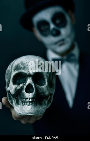 beängstigend Porträt eines jungen Mannes mit mexikanischen Calaveras Make-up trägt Hut und Fliege um den Hals und hält einen gruseligen Schädel vor h Stockfoto