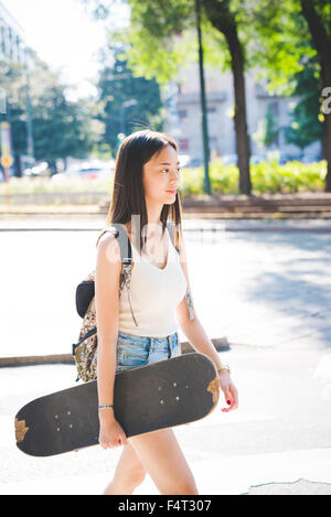 Knie-Figur des jungen hübschen asiatischen lange braune glatte Haare Frau Skater hält eine Skateboard zu Fuß in der Stadt im Freien, mit Blick auf Links, lächelnd - sportlich, Spaß, Glück-Konzept Stockfoto