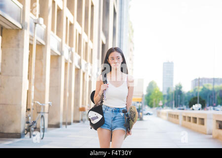 Knie-Figur des jungen hübschen asiatischen Langhaar Frau Skater zu Fuß in der Stadt mit Blick auf Links, nachdenklich – denken, Zukunft, entspannende Konzept Stockfoto