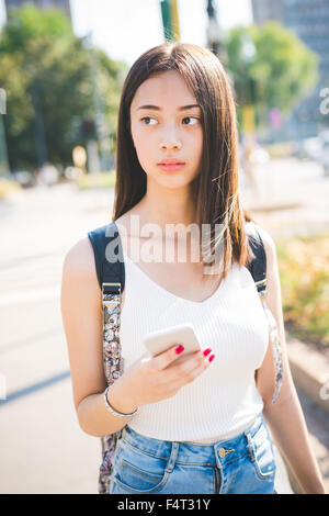 Knie-Figur des jungen schönen asiatischen lange braune glatte Haare Frau hält und per Smartphone, mit Blick auf rechts - Technologie, soziales Netzwerk, Kommunikations-Konzept Stockfoto